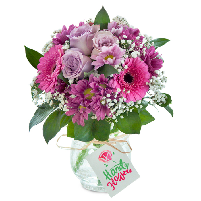 Capturing the lively spirit of Parsons Green, this mixed bouquet bursts forth with saturated shades of pink, purple, and lavender, displayed imaginatively within a textured clear glass vase set against a luminous white background. At the heart, elegant lavender roses slowly unfurl, surrounded by a joyful circle of large hot pink Gerbera daisies, whose bold faces and deep-dyed centres draw the eye. Smaller magenta chrysanthemum-like blooms cluster around, their intricate petals and subtle green-yellow centres creating depth and a tapestry of visual interest. Airy baby's breath threads through the design, offering pockets of softness that lighten the composition, much like spring clouds drifting above Hurlingham Park. Brilliant green foliage peeks generously from beneath the blooms, grounding the palette and enhancing the bouquet's fullness. The vase itself is finished with a understated raffia ribbon and a small, simple white gift tag awaiting a personal wish. This beautifully crafted arrangement, echoing the care and creativity of Parsons Green's local florists, is perfectly suited as a birthday surprise, a token of gratitude, or a charming centrepiece for a family gathering-each bloom promising the highest standard of floral artistry and freshness for delivery in SW6.